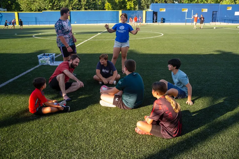 Niños y niñas de la escuela sentados en corro escuchan a su entrenador en el campo de Gines